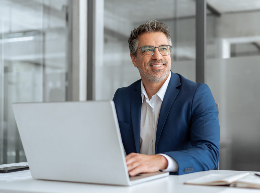 Middle age latin male entrepreneur businessman working on financial sale work at desk in office. Mature indian business man in suit using pc laptop for trading, looking smiling, dreaming at copy space